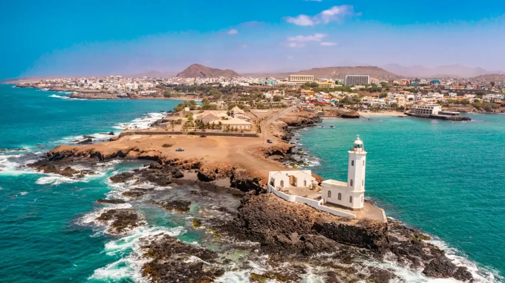 A scenic view of a coastal city, reminiscent of Praia, with a lighthouse perched on a rocky outcrop surrounded by turquoise ocean waters. The city stretches across the background with mountains silhouetted under a clear blue sky.