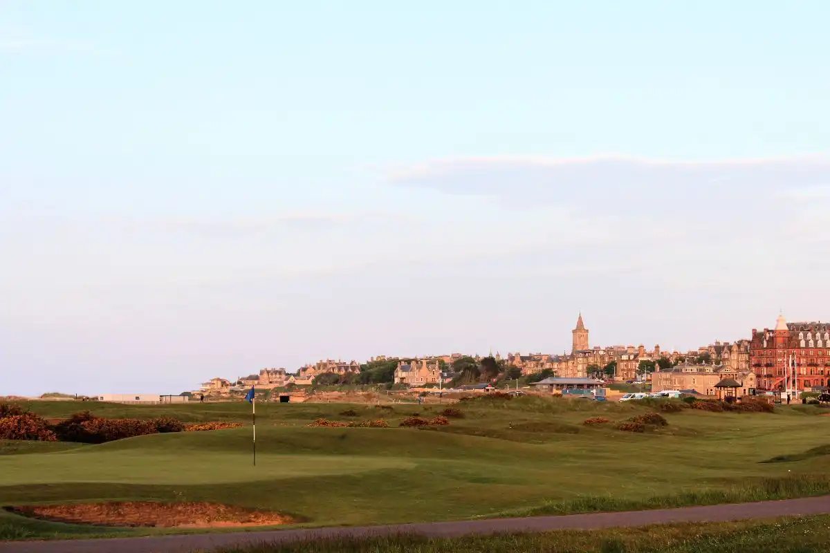 A panoramic view showcases a lush golf course in the foreground, set against the backdrop of a historic town and its prominent clock tower, all beneath a vast expanse of clear blue sky.