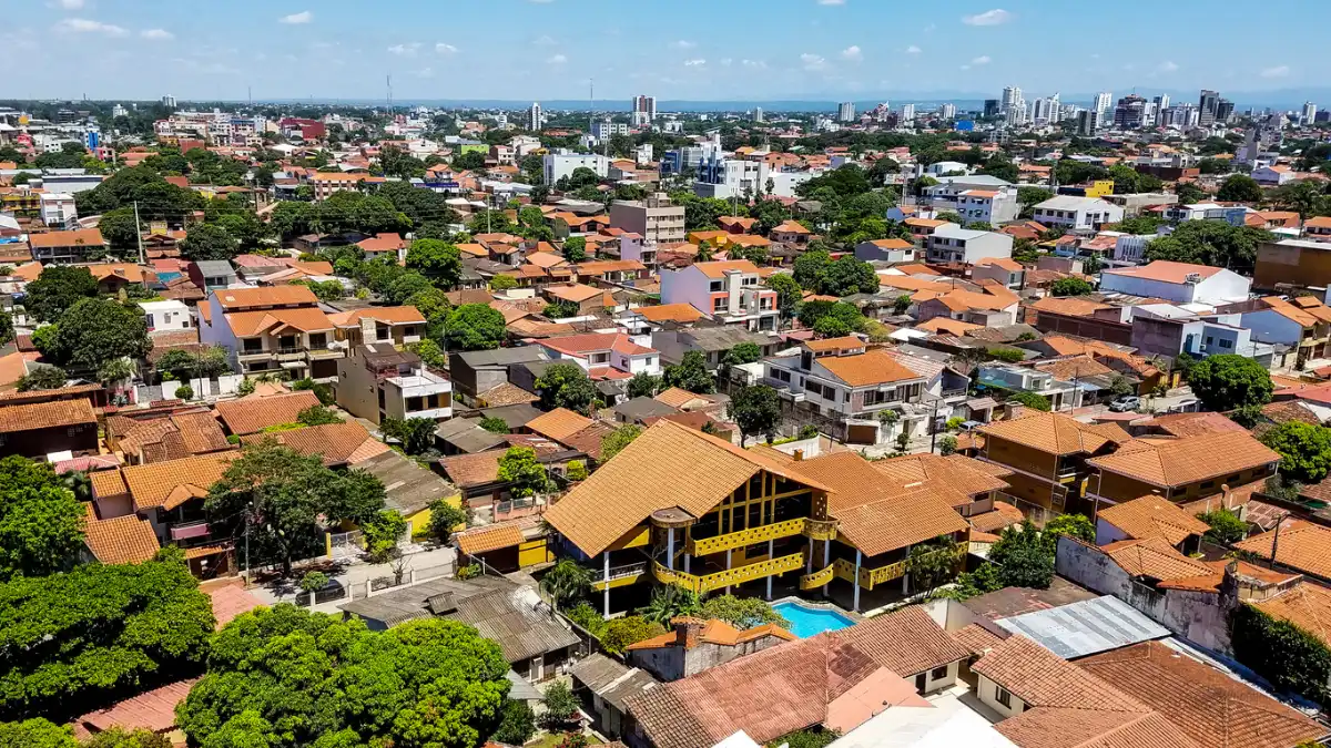 Aerial view of Santa Cruz de la Sierra, showcasing densely packed red-roofed buildings surrounded by lush green trees. A large building with a swimming pool stands out in the center, while the skyline features a mix of low-rise and a few taller structures under a blue sky.