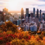 A vibrant cityscape at sunset with Montreal's iconic skyscrapers in the background. In the foreground, autumn foliage in shades of red, orange, and yellow creates a colorful contrast. The sky is partly cloudy with the sun casting a warm glow over the scene.