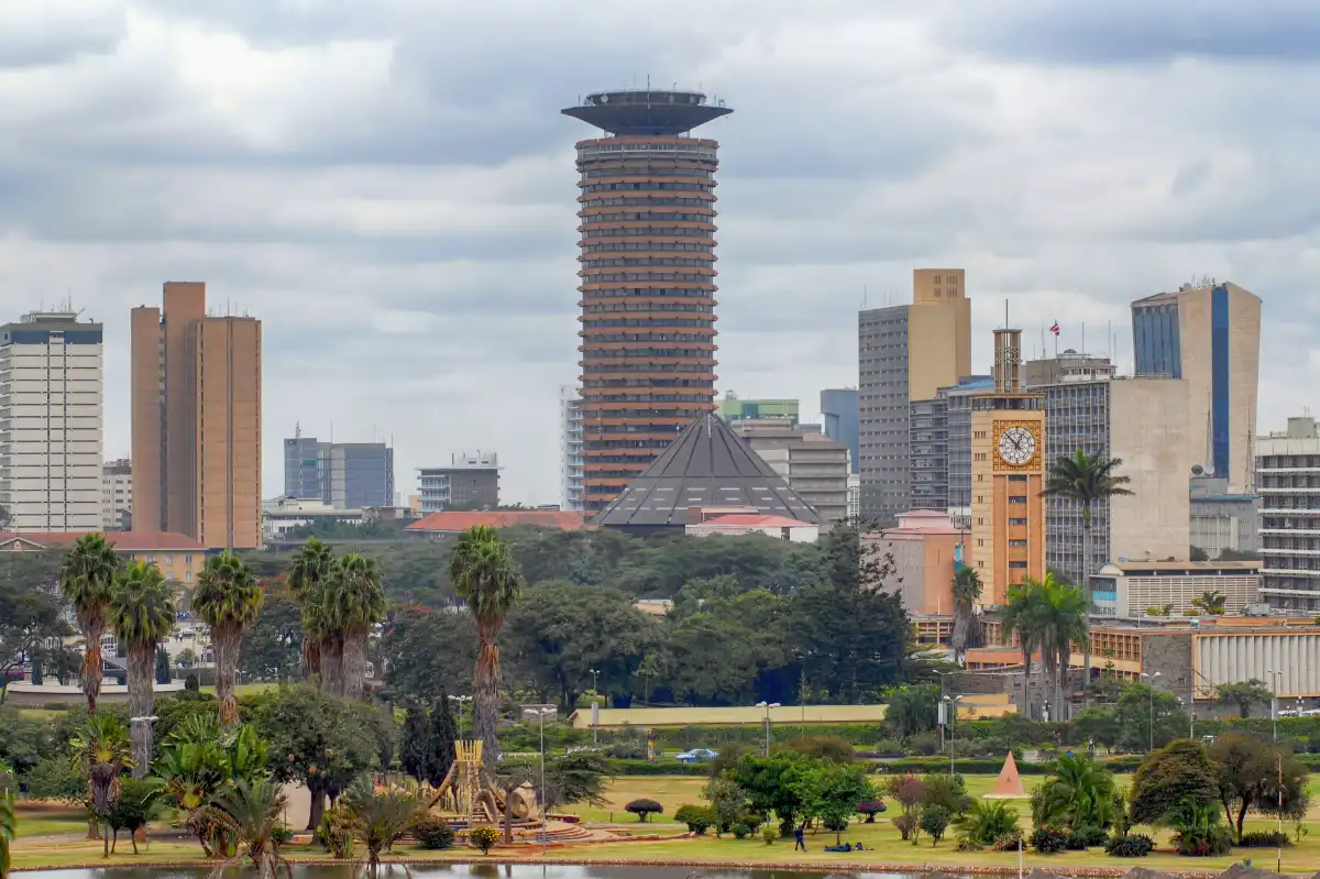 The Nairobi skyline showcases the distinctive cylindrical Kenyatta International Convention Centre, surrounded by modern buildings and lush greenery, all set under a dramatic cloudy sky.