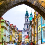 A picturesque street scene in Prague, framed by a stone arch, reveals colorful historic buildings. A prominent church tower with a green dome rises in the background under a clear sky, capturing the charm of this European city.