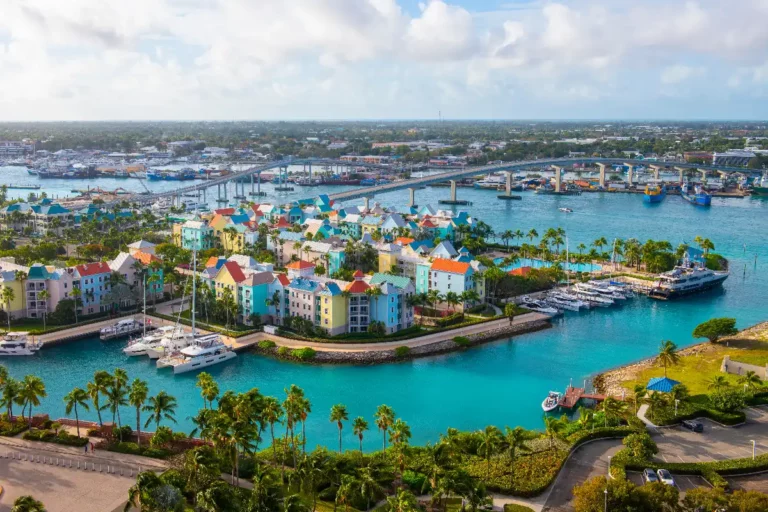 Aerial view of a tropical island resort near Nassau, featuring colorful buildings surrounded by turquoise water and yachts. A bridge spans the water, connecting to a bustling cityscape in the background under a partly cloudy sky.