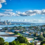 Auckland's scenic view showcases a mix of urban skyline and residential areas, where the iconic Sky Tower rises amidst city buildings. Lush greenery and the tranquil harbor beautifully complement this Auckland landscape under a partly cloudy sky.