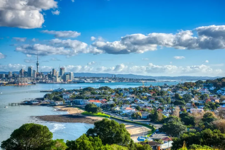 Auckland's scenic view showcases a mix of urban skyline and residential areas, where the iconic Sky Tower rises amidst city buildings. Lush greenery and the tranquil harbor beautifully complement this Auckland landscape under a partly cloudy sky.