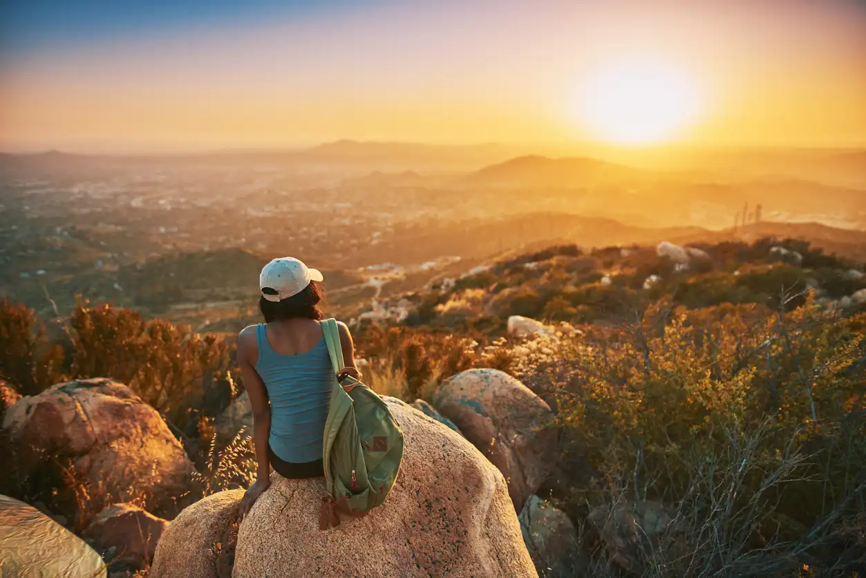 A person wearing a cap and carrying a backpack sits on a rock, overlooking a scenic West Coast sunset from a hilltop. The landscape features rolling hills, distant mountains, and cityscape bathed in warm, golden light.