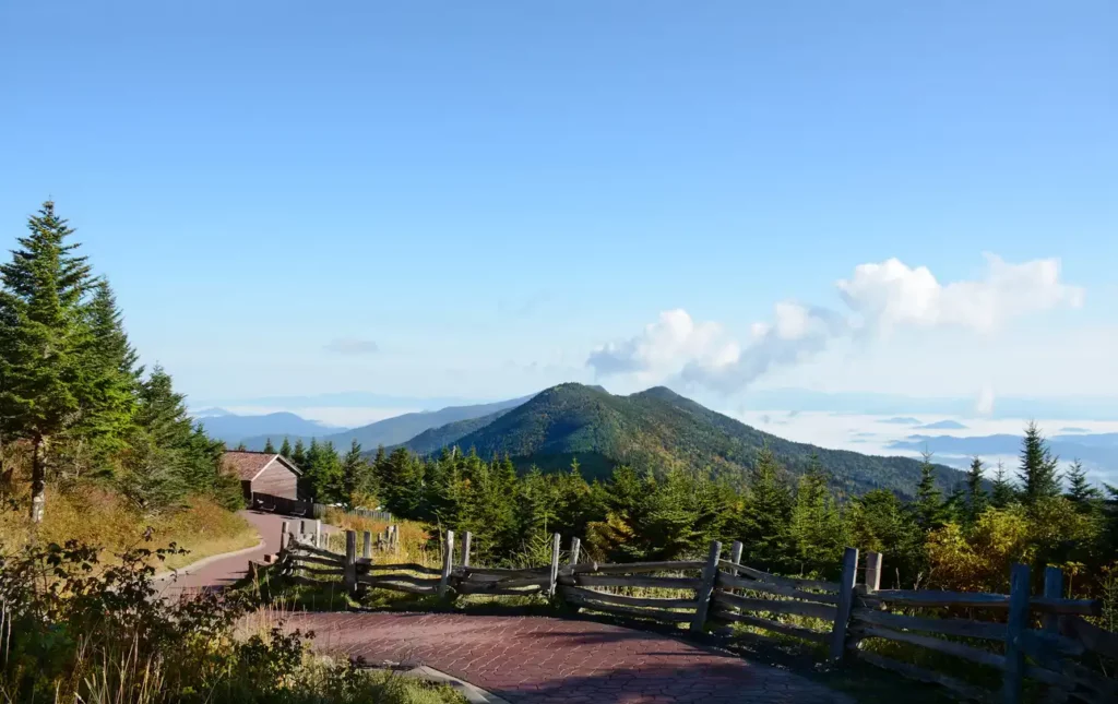 A paved path with a wooden fence leads to a small building on a hillside, surrounded by trees. In the distance, the majestic Appalachian Mountains are visible under a clear blue sky with a few scattered clouds.
