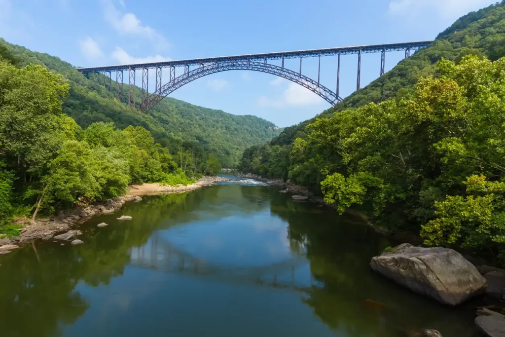 A wide-angle view of a lush, green valley with a large arched bridge spanning across a river. The river reflects the clear blue sky and surrounding trees. Forested hills frame the scene in the heart of the Appalachian Mountains, creating a tranquil and picturesque landscape.