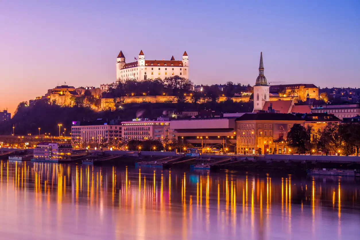 A scenic view of Bratislava, Slovakia, at dusk. The illuminated Bratislava Castle sits prominently on a hill, with reflections shimmering in the calm river below. The skyline includes the spire of St. Martin's Cathedral, all beautifully enhanced by twilight colors.